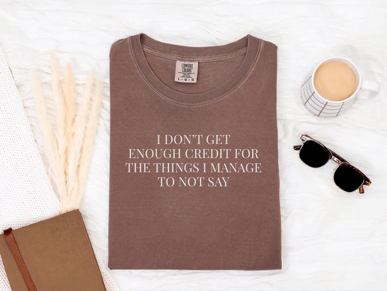May include: A brown t-shirt with the text "I DON'T GET ENOUGH CREDIT FOR THE THINGS I MANAGE TO NOT SAY". The shirt is laid flat on a white surface, with a cup of coffee, sunglasses, and a notebook nearby.