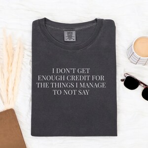 May include: Dark gray t-shirt with the text "I DON'T GET ENOUGH CREDIT FOR THE THINGS I MANAGE TO NOT SAY" in white. The shirt is folded and surrounded by a coffee mug, sunglasses, and a notebook.