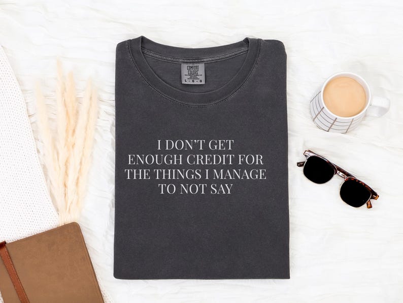 May include: Dark gray t-shirt with the text "I DON'T GET ENOUGH CREDIT FOR THE THINGS I MANAGE TO NOT SAY" in white. The shirt is folded and surrounded by a coffee mug, sunglasses, and a notebook.