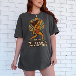 May include: Dark gray t-shirt featuring a graphic of a woman lifting weights, with the text "ALL THE PRETTY GIRLS WALK LIKE THIS". The design includes orange, yellow, and green accents, creating a retro aesthetic. The shirt is a comfortable, casual wear.