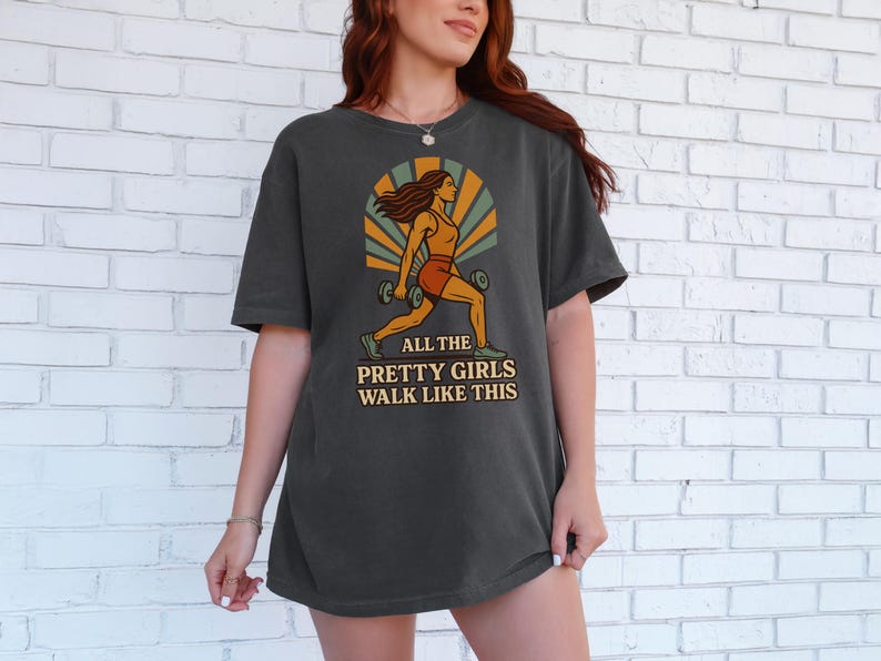 May include: Dark gray t-shirt featuring a graphic of a woman lifting weights, with the text "ALL THE PRETTY GIRLS WALK LIKE THIS". The design includes orange, yellow, and green accents, creating a retro aesthetic. The shirt is a comfortable, casual wear.