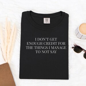 May include: Black t-shirt with the quote "I DON'T GET ENOUGH CREDIT FOR THE THINGS I MANAGE TO NOT SAY" in white. The shirt is on a white surface with a cup of coffee, sunglasses, and a notebook.