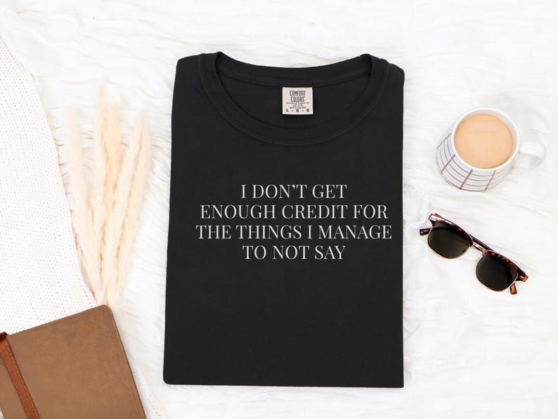 May include: Black t-shirt with the quote "I DON'T GET ENOUGH CREDIT FOR THE THINGS I MANAGE TO NOT SAY" in white. The shirt is on a white surface with a cup of coffee, sunglasses, and a notebook.