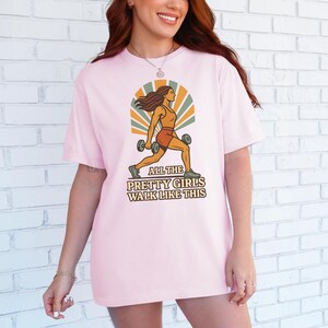 May include: Light pink t-shirt with a retro graphic of a woman lifting dumbbells, set against a sunburst design. The text "ALL THE PRETTY GIRLS WALK LIKE THIS" is below the image. The shirt is a casual, oversized fit.