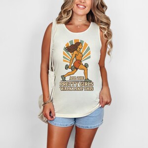 May include: Cream-colored tank top with a retro illustration of a woman lifting weights, set against a sunburst design. The text reads "ALL THE PRETTY GIRLS WALK LIKE THIS". The shirt is paired with denim shorts.