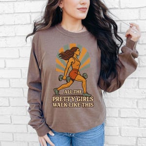 May include: Brown long-sleeve shirt with a graphic of a woman lifting weights. The graphic is in orange, green, and brown tones, with the text "ALL THE PRETTY GIRLS WALK LIKE THIS". The shirt is worn with blue jeans.
