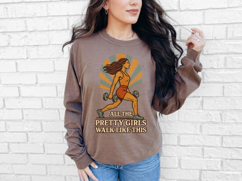 May include: Brown long-sleeve shirt with a graphic of a woman lifting weights. The graphic is in orange, green, and brown tones, with the text "ALL THE PRETTY GIRLS WALK LIKE THIS". The shirt is worn with blue jeans.