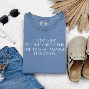 May include: A light blue t-shirt with the text "I DON'T GET ENOUGH CREDIT FOR THE THINGS I MANAGE TO NOT SAY". The image also includes denim shorts, sunglasses, a necklace, and beige clogs.