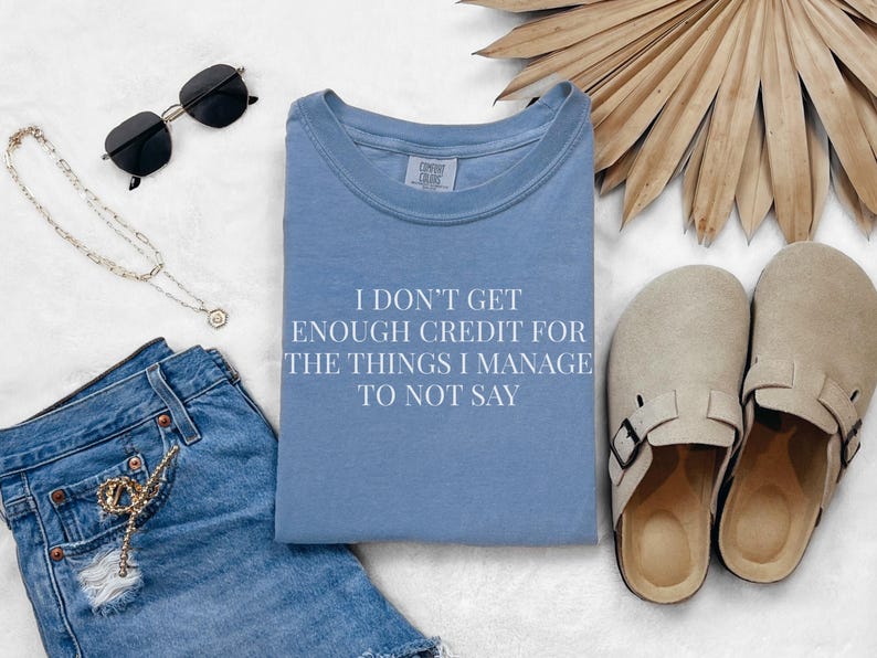May include: A light blue t-shirt with the text "I DON'T GET ENOUGH CREDIT FOR THE THINGS I MANAGE TO NOT SAY". The image also includes denim shorts, sunglasses, a necklace, and beige clogs.