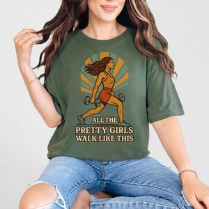 May include: Olive green t-shirt with a graphic of a woman lifting weights, set against a retro sunburst design. The text "ALL THE PRETTY GIRLS WALK LIKE THIS" is printed below the illustration. The shirt is worn by a person.