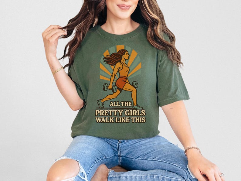 May include: Olive green t-shirt with a graphic of a woman lifting weights, set against a retro sunburst design. The text "ALL THE PRETTY GIRLS WALK LIKE THIS" is printed below the illustration. The shirt is worn by a person.