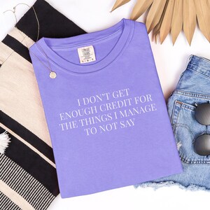 May include: A lilac-colored t-shirt with the text "I DON'T GET ENOUGH CREDIT FOR THE THINGS I MANAGE TO NOT SAY" in white. The shirt is paired with denim shorts, sunglasses, a tote bag, and a necklace.