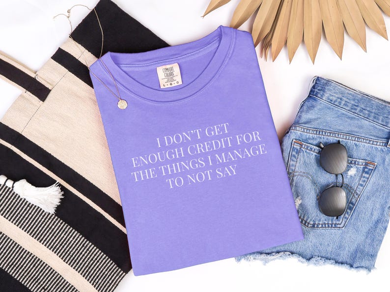 May include: A lilac-colored t-shirt with the text "I DON'T GET ENOUGH CREDIT FOR THE THINGS I MANAGE TO NOT SAY" in white. The shirt is paired with denim shorts, sunglasses, a tote bag, and a necklace.