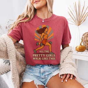 May include: A maroon t-shirt with a graphic of a woman lifting weights, set against a retro sunburst design. The text reads "ALL THE PRETTY GIRLS WALK LIKE THIS". The shirt is paired with denim shorts.