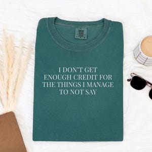 May include: A teal-colored t-shirt with the text "I DON'T GET ENOUGH CREDIT FOR THE THINGS I MANAGE TO NOT SAY" in white. The shirt is on a white surface with a cup of coffee and sunglasses.