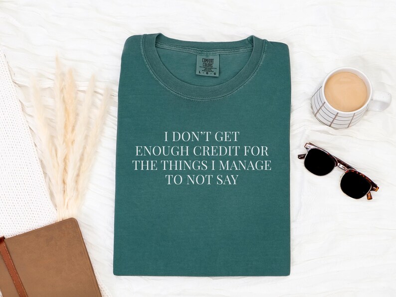 May include: A teal-colored t-shirt with the text "I DON'T GET ENOUGH CREDIT FOR THE THINGS I MANAGE TO NOT SAY" in white. The shirt is on a white surface with a cup of coffee and sunglasses.