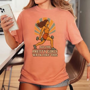 May include: Peach-colored t-shirt featuring a retro illustration of a woman exercising with dumbbells. The graphic includes the text "ALL THE PRETTY GIRLS WALK LIKE THIS". The shirt is a comfortable, casual style.