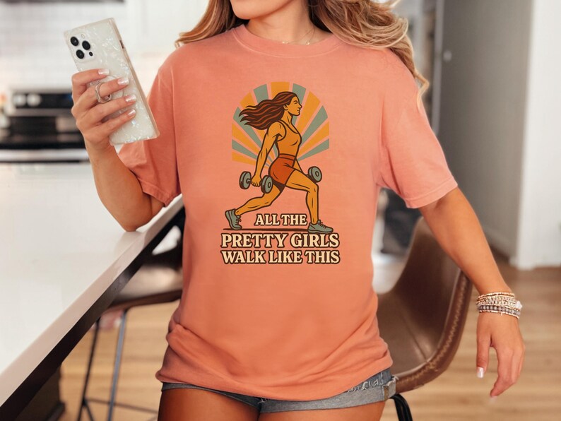 May include: Peach-colored t-shirt featuring a retro illustration of a woman exercising with dumbbells. The graphic includes the text "ALL THE PRETTY GIRLS WALK LIKE THIS". The shirt is a comfortable, casual style.