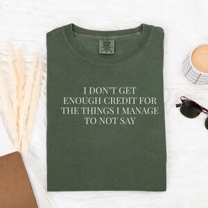 May include: Olive green t-shirt with the text "I DON'T GET ENOUGH CREDIT FOR THE THINGS I MANAGE TO NOT SAY". The shirt is laid flat on a white surface, next to a coffee mug, sunglasses, and a notebook.