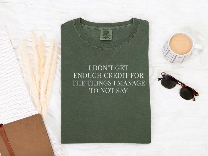 May include: Olive green t-shirt with the text "I DON'T GET ENOUGH CREDIT FOR THE THINGS I MANAGE TO NOT SAY". The shirt is laid flat on a white surface, next to a coffee mug, sunglasses, and a notebook.