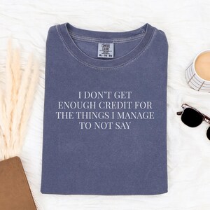 May include: A navy blue t-shirt with the text "I DON'T GET ENOUGH CREDIT FOR THE THINGS I MANAGE TO NOT SAY" in white. The shirt is surrounded by a coffee cup, sunglasses, and a notebook.
