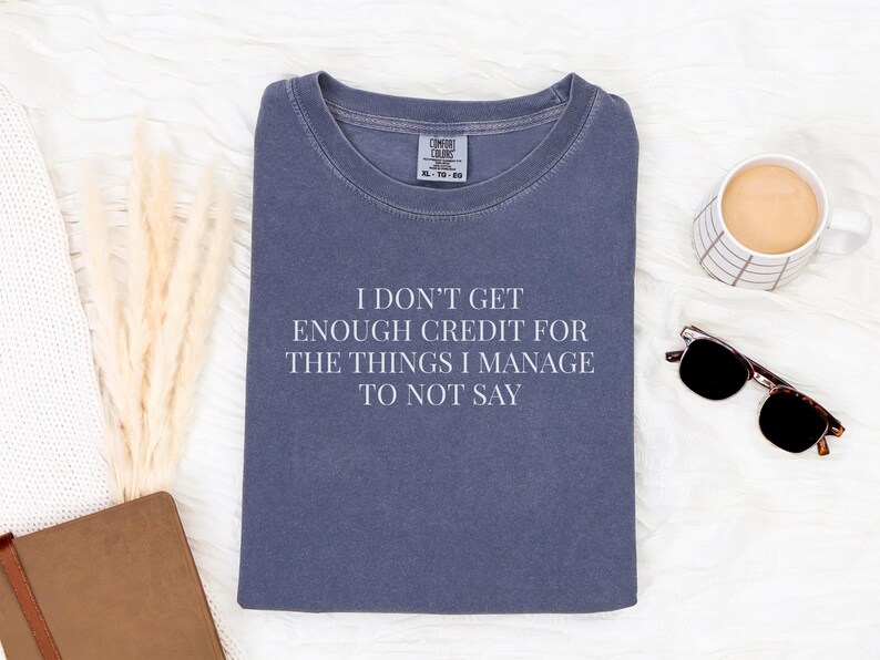 May include: A navy blue t-shirt with the text "I DON'T GET ENOUGH CREDIT FOR THE THINGS I MANAGE TO NOT SAY" in white. The shirt is surrounded by a coffee cup, sunglasses, and a notebook.