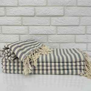 Può includere: Un mucchio di asciugamani a righe grigie e bianche con frange sui bordi. Gli asciugamani sono piegati e disposti su una superficie bianca.