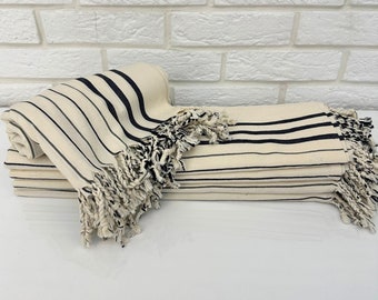 May include: Several folded cream-colored towels with black stripes and fringed edges are stacked. One towel is rolled up, displaying the striped pattern. The towels are arranged on a white surface against a white brick wall.