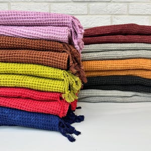 Pode incluir: Uma pilha de toalhas de tecido waffle em várias cores, incluindo roxo, marrom, verde, vermelho, azul e cinza. As toalhas têm borlas nas pontas.