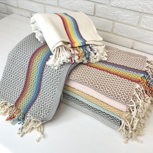 May include: A stack of colorful Turkish towels with diamond patterns and fringed edges. One towel is folded, displaying a rainbow stripe. The towels come in various colors, including gray, brown, pink, and blue, and are made of soft, absorbent material.