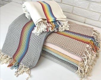 May include: A stack of colorful Turkish towels with diamond patterns and fringed edges. One towel is folded, displaying a rainbow stripe. The towels come in various colors, including gray, brown, pink, and blue, and are made of soft, absorbent material.