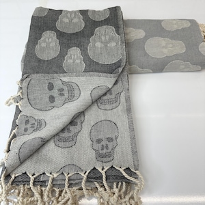 May include: A folded grey and black skull-patterned towel with cream-coloured fringe. The towel features a repeating pattern of skulls in a lighter grey colour. The towel is folded over another towel with a similar skull pattern.