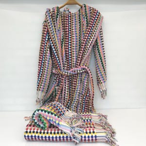 Puede incluir: Un albornoz estampado multicolor y una toalla plegada a juego. El albornoz, colgado de una percha de madera, presenta un estampado de puntos en varios colores, como rosa, azul y rojo. La toalla tiene un diseño similar y flecos.