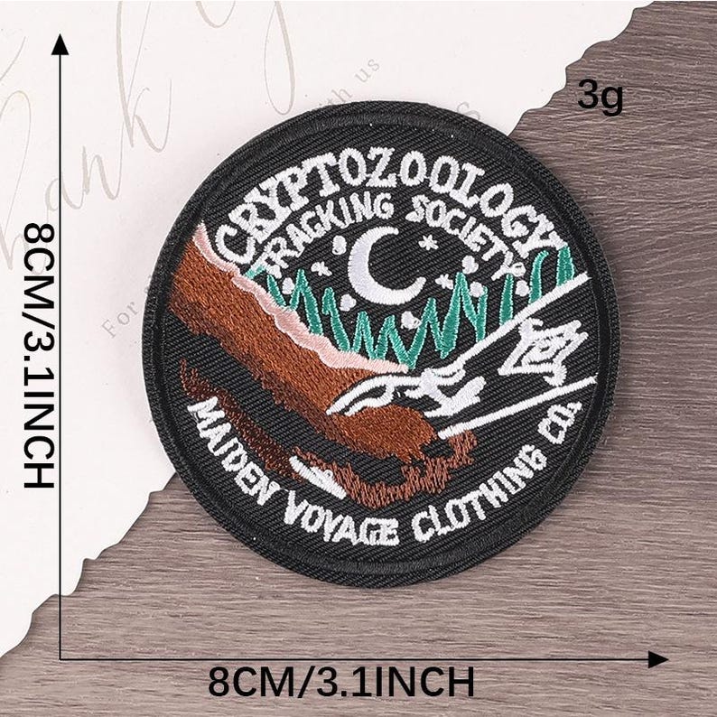 Peut inclure: Un &eacute;cusson rond brod&eacute; noir avec le texte "CRYPTOZOOLOGY TRACKING SOCIETY" et "MAIDEN VOYAGE CLOTHING CO." Le motif comprend une montagne marron, des arbres verts, un croissant de lune et du texte blanc. Il mesure 8 cm de diam&egrave;tre.