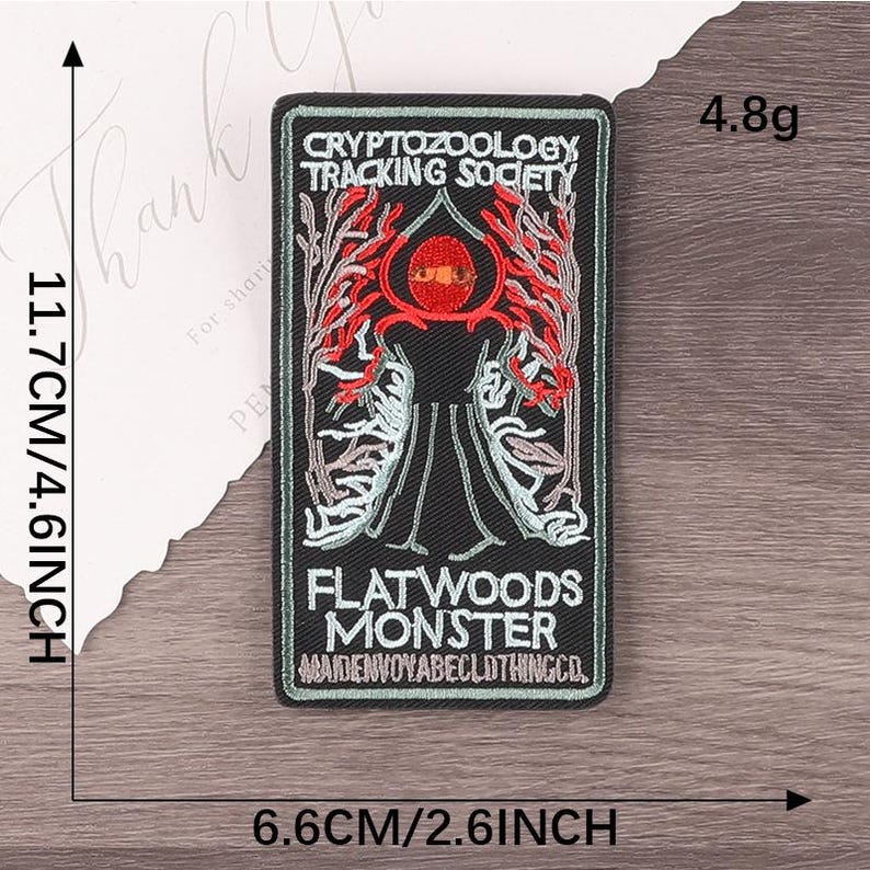 Peut inclure: &Eacute;cusson rectangulaire noir avec le texte "CRYPTOZOOLOGY TRACKING SOCIETY" et "FLATWOODS MONSTER". Le patch pr&eacute;sente un motif rouge et noir avec une figure centrale et mesure 11,7 cm sur 6,6 cm.