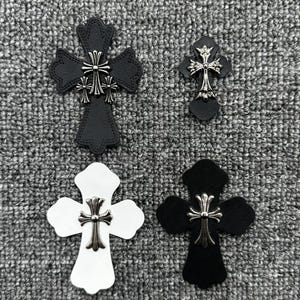 May include: Four decorative cross-shaped embellishments in black and white, with silver-colored metal accents. The crosses vary in size and design, with intricate detailing on the metalwork. The background is a textured gray surface.