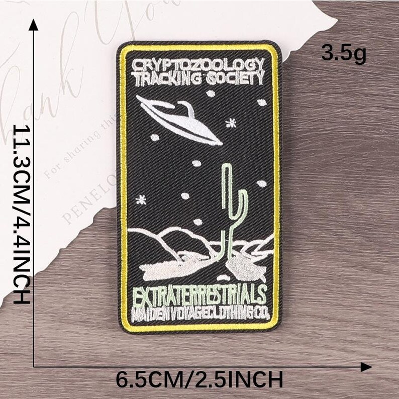 Peut inclure: Un &eacute;cusson rectangulaire noir avec une bordure jaune, pr&eacute;sentant du texte et des illustrations blanches. L'&eacute;cusson indique "CRYPTOZOOLOGY TRACKING SOCIETY" et "EXTRATERRESTRIALS MAIDEN VOYAGE CLOTHING CO." Il repr&eacute;sente un OVNI, un cactus et des &eacute;toiles. L'&eacute;cusson mesure 11,3 cm sur 6,5 cm et p&egrave;se 3,5 g.