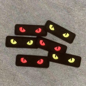 Könnte beinhalten: Eine Sammlung schwarzer rechteckiger Aufnäher, jeder mit einem Paar leuchtender Katzenaugen bestickt. Die Augen sind entweder leuchtend rot oder neongelb und bilden einen auffälligen Kontrast zum dunklen Hintergrund. Diese Aufnäher sind wahrscheinlich für Kleidung oder Accessoires.
