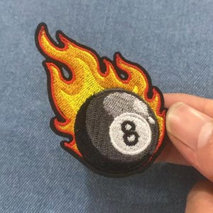 Puede incluir: Parche bordado con una bola ocho negra envuelta en llamas. El parche tiene un borde negro y está detallado con hilo amarillo, naranja y rojo. El número 8 es blanco.