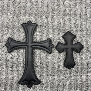 Puede incluir: Dos cruces de cuero negro, una grande y otra pequeña, se muestran sobre un fondo gris texturizado. Las cruces tienen un diseño de borde decorativo y puntiagudo y detalles cosidos, lo que sugiere un parche o aplique.