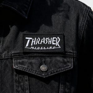 Può includere: Giacca di jeans nera con tasca sul petto e bottoni argentati. La giacca presenta una toppa ricamata bianca con la scritta "Thrasher Magazine". Un capo di abbigliamento esterno classico e casual.