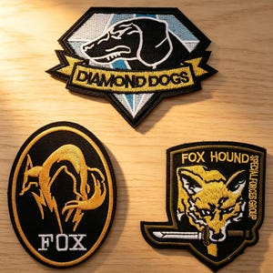 May include: Three embroidered patches with military-style designs. The top patch features a black dog within a diamond shape and the words "DIAMOND DOGS." The other patches have a fox design and the words "FOX" and "FOX HOUND SPECIAL FORCES GROUP."