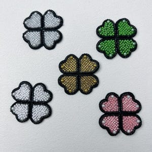 以下が含まれることがあります： 銀、緑、金、白、ピンクのビーズで飾られた四つ葉のクローバーのパッチが5つ。各クローバーは黒い縁取りがあり、小さなビーズと真珠で飾られています。クローバーは白い表面に配置されています。