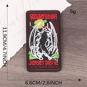 Peut inclure: Un &eacute;cusson rectangulaire avec le texte "CRYPTOZOOLOGY TRACKING SOCIETY" et "JERSEY DEVIL". L'&eacute;cusson pr&eacute;sente une illustration en noir et blanc d'une cr&eacute;ature ail&eacute;e aux yeux rouges. L'&eacute;cusson mesure 11,9 cm sur 6,6 cm.