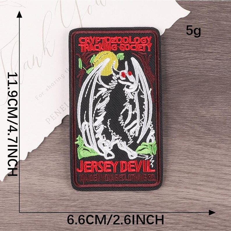 Peut inclure: Un &eacute;cusson rectangulaire avec le texte "CRYPTOZOOLOGY TRACKING SOCIETY" et "JERSEY DEVIL". L'&eacute;cusson pr&eacute;sente une illustration en noir et blanc d'une cr&eacute;ature ail&eacute;e aux yeux rouges. L'&eacute;cusson mesure 11,9 cm sur 6,6 cm.