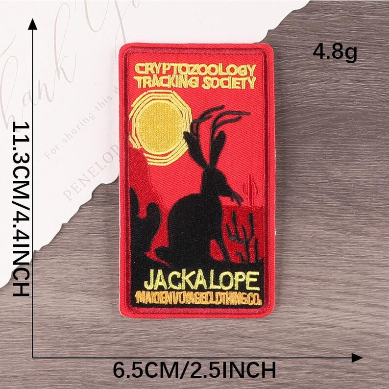 Peut inclure: &Eacute;cusson rectangulaire rouge avec un jackalope noir, un soleil jaune et des cactus. Le texte indique "CRYPTOZOOLOGY TRACKING SOCIETY" et "JACKALOPE". Le patch mesure 11,3 cm de haut et 6,5 cm de large.