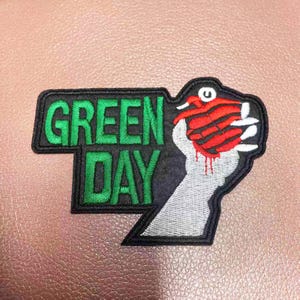 Puede incluir: Parche bordado con el logotipo de la banda Green Day sobre fondo negro, texto verde y una mano blanca sosteniendo un corazón rojo con contorno negro.