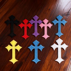 Peut inclure: Plusieurs croix brodées de différentes couleurs, dont noir, rouge, violet, rose, bleu, jaune et blanc. Les croix sont disposées sur une surface en bois foncé, mettant en valeur leurs coutures détaillées et leurs teintes vives.