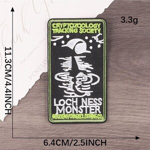 Peut inclure: Un &eacute;cusson rectangulaire avec un fond noir et une bordure verte. L'&eacute;cusson comporte un texte blanc qui dit "CRYPTOZOOLOGY TRACKING SOCIETY" et "LOCH NESS MONSTER". L'&eacute;cusson mesure 11,3 cm de haut et 6,4 cm de large.