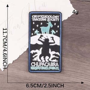 Peut inclure: Un &eacute;cusson rectangulaire avec un fond noir et une bordure bleue. L'&eacute;cusson pr&eacute;sente une ch&egrave;vre turquoise et le texte "CRYPTOZOOLOGY TRACKING SOCIETY" et "CHUPACABRA". L'&eacute;cusson mesure 11,7 cm de haut et 6,5 cm de large.